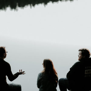 three people sitting at the shore of a lake